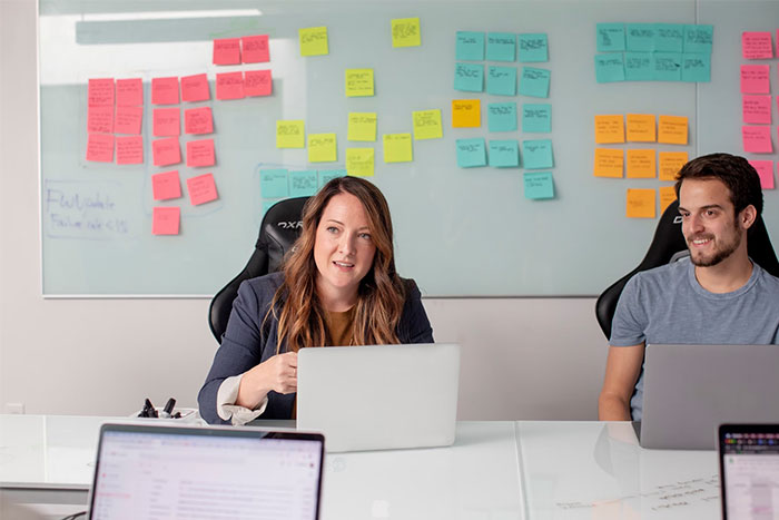 Two professionals in a conference room discussing work, whiteboard covered with sticky notes, laptops open on the table.