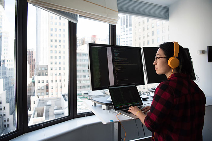 Person coding at a desk with multiple screens, wearing headphones, in a city office environment.