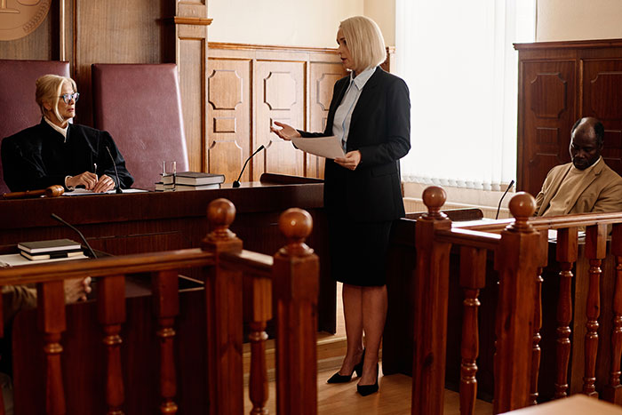 Woman in a courtroom setting discussing a family feud over a heirloom, standing before a judge and a seated man.