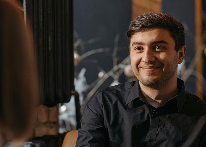 A man in a black shirt smiling warmly in a cozy, dimly lit setting, conveying a unique and different charm.