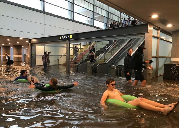 Flooded airport terminal showing people floating on inflatable rings, highlighting nature disaster resilience.