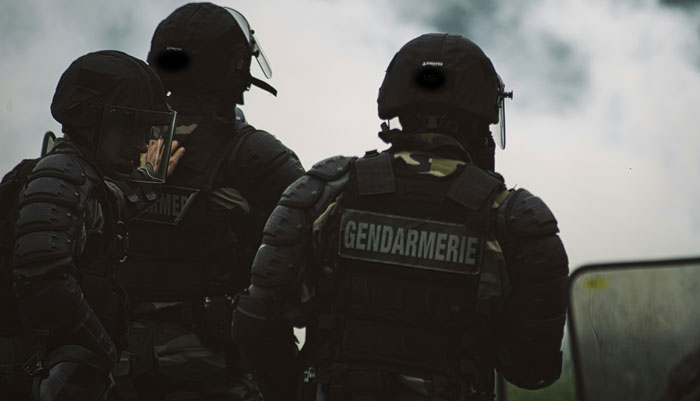 Gendarmerie officers in tactical gear, standing together amidst smoke.