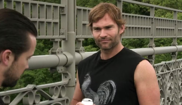 Man in a sleeveless black shirt with a rooster design on a bridge, holding a can. Celebrities and fame theme.