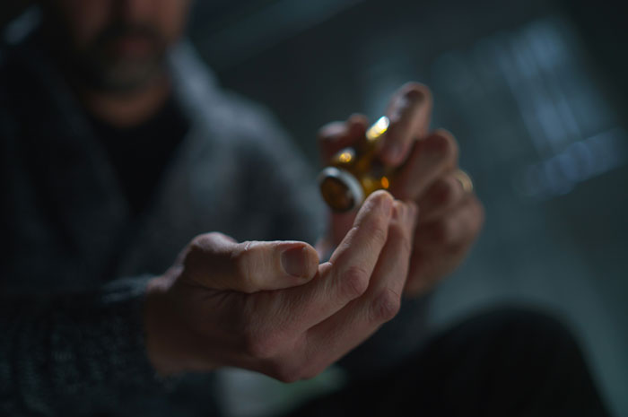 Man in a dark setting pouring pills from a bottle into his hand, symbolizing sacrifice and survival stories shared.
