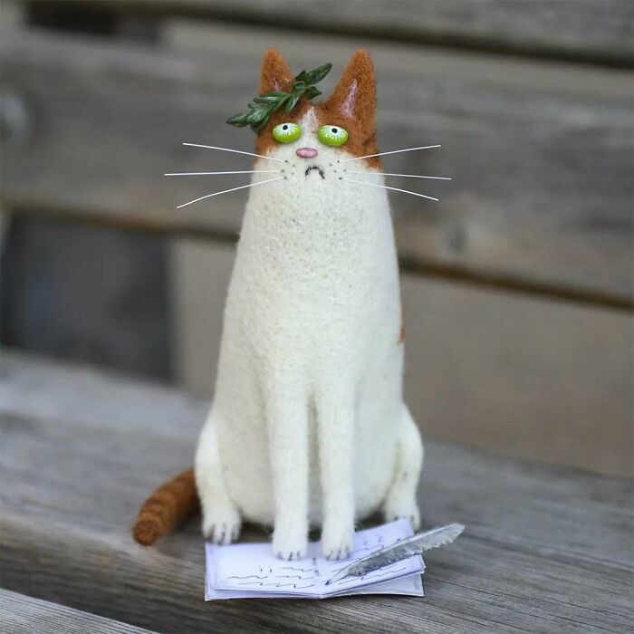 Strange art doll of a funny-looking cat with green eyes, wearing a leaf crown, sitting on papers with a pen nearby.