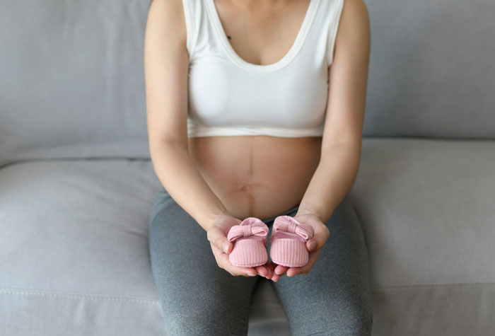 Pregnant woman sitting on a couch holding pink baby shoes, symbolizing people who survived due to another’s sacrifice.