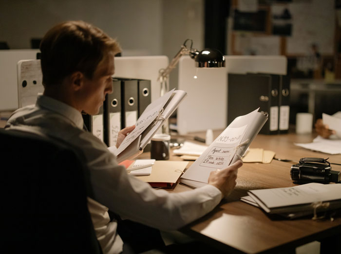 Man reviewing documents in a dimly lit office, reflecting on stories of survival and sacrifice shared by others.