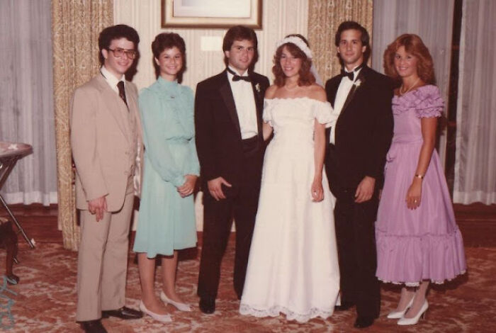 Group in 80s fashion trends with suits, pastel dresses, and ruffles, posing at an indoor event.