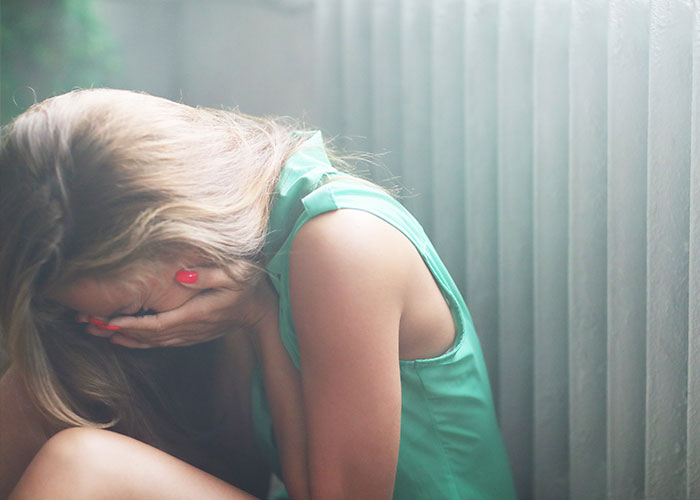 Woman in green dress sitting by a wall, covering her face with her hands, appearing upset and emotional.