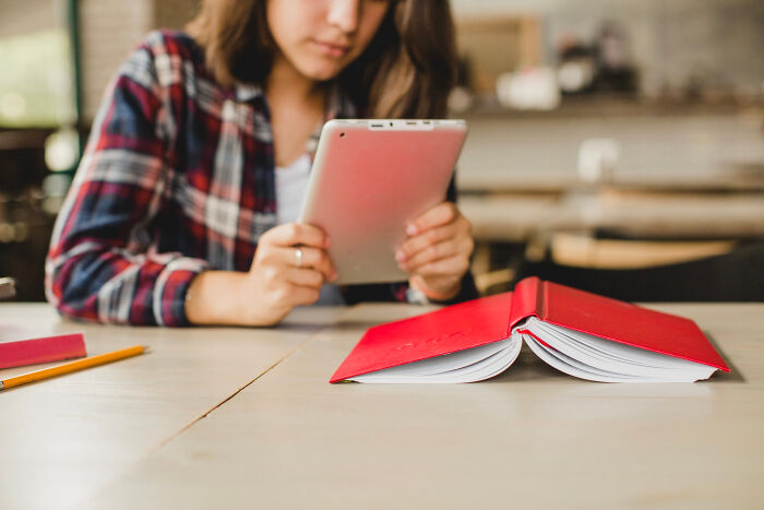 Person using modern technology with a tablet at a table, having an open book and pencil in front of them.
