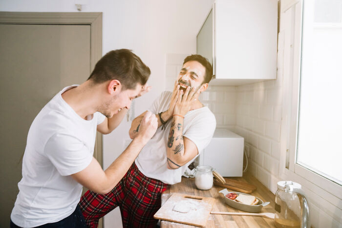 Two men in a kitchen playfully applying flour to each other's faces, surrounded by baking ingredients, illustrating home rules.