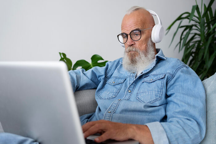 Older man using a laptop, wearing headphones and glasses, illustrating modern technology engagement challenges.