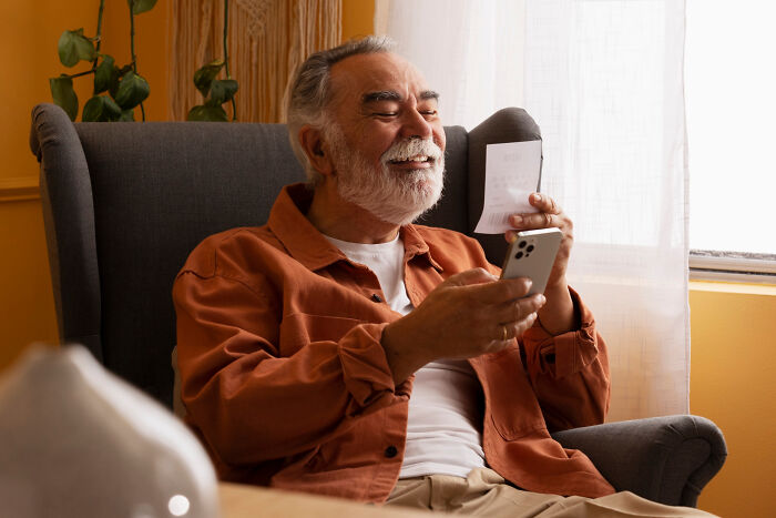 Older man smiling, sitting in a chair holding smartphone, depicting modern technology use among older adults.
