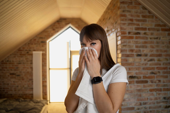Person in a brick-walled room adhering to strict home rules, holding a cloth near their face, wearing a smartwatch.