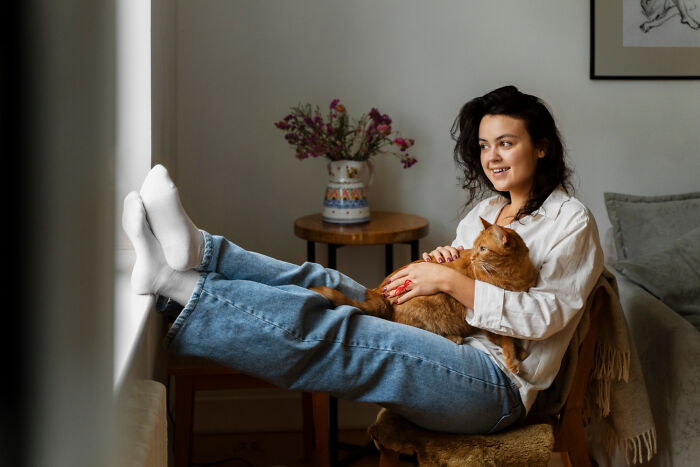 Woman relaxing at home, wearing jeans and a white shirt, holding a cat, surrounded by cozy decor, highlighting strict home rules.