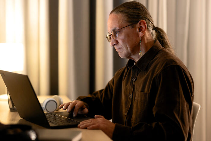 Older person with a ponytail using modern technology, focused on a laptop in a dimly lit room.