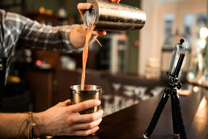 Bartender pouring drink into shaker, with smartphone on tripod recording the process.
