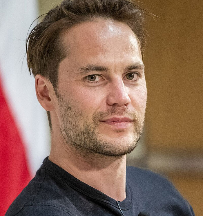 Unknown person with short dark hair and a beard, wearing a black shirt, gazing toward the camera.