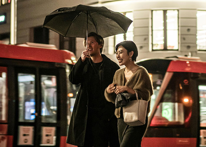 A man holding an umbrella over a woman, both smiling, walking past a city street with buses, showing he's different.