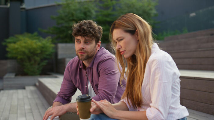 A woman sitting with a man, looking concerned, holding a coffee cup outdoors.