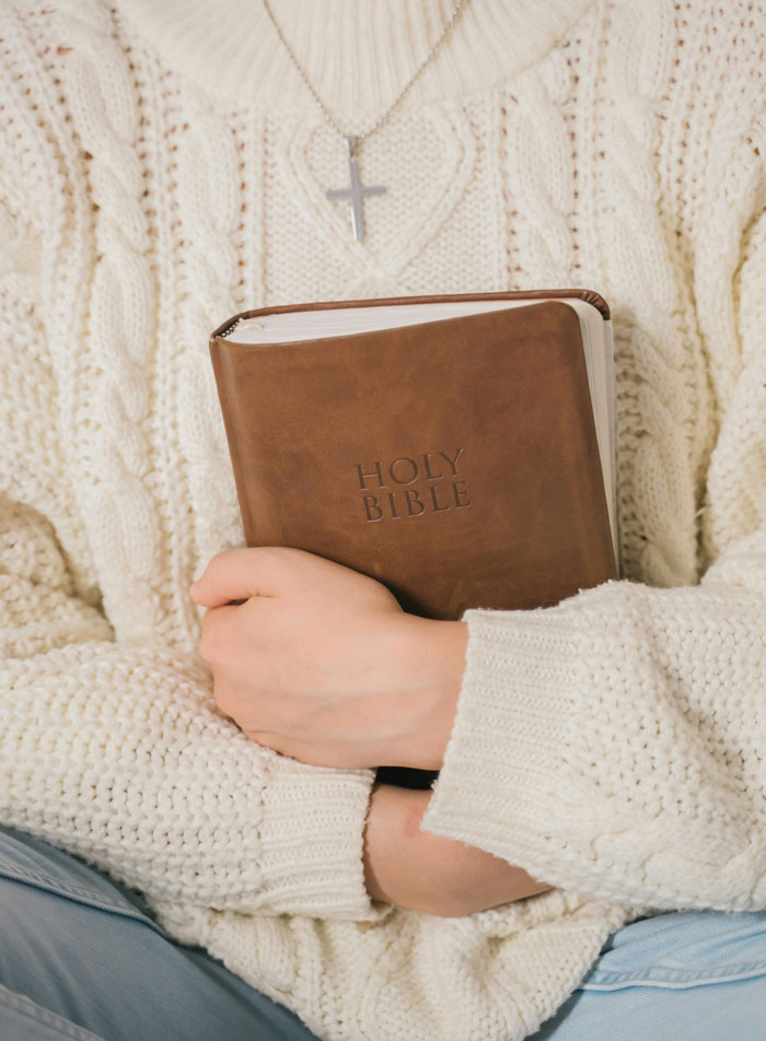 Person holding a Bible, wearing a cream sweater and cross necklace, symbolizing family secrets.