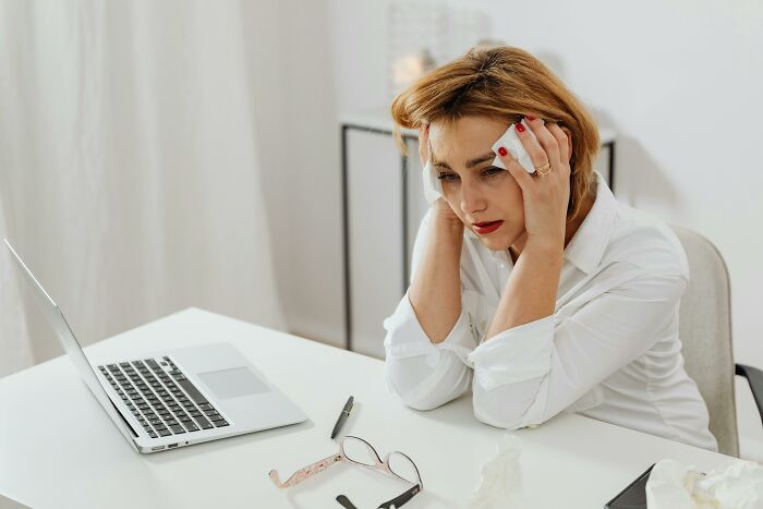 Woman in a white blouse at a desk, stressed, contemplating consequences, with a laptop and glasses nearby.