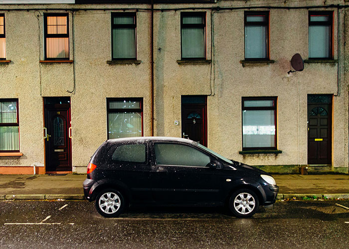 A black car parked on a quiet street, with apartment buildings in the background, highlighting unique urban moments.