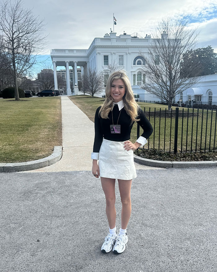 White House correspondent in a black top and white skirt, standing outside the White House, facing criticism for outfit choice.