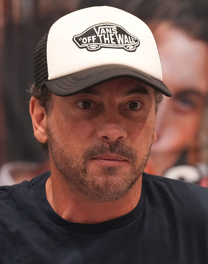 Celebrity wearing cap and black shirt, with beard, in a close-up at an event.