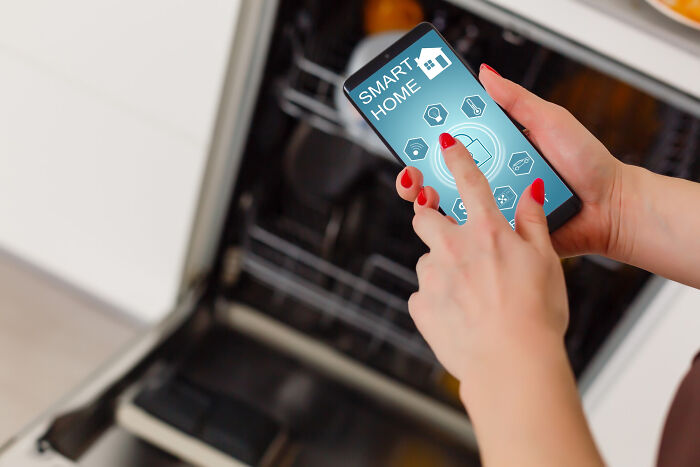 Person using smartphone to control smart home technology, near a dishwasher.