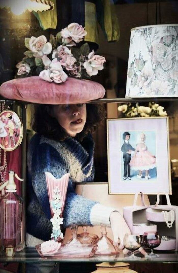 A woman arranges shop window decor under pastel floral hat; a moment through Nadia Eeckhout’s lens.