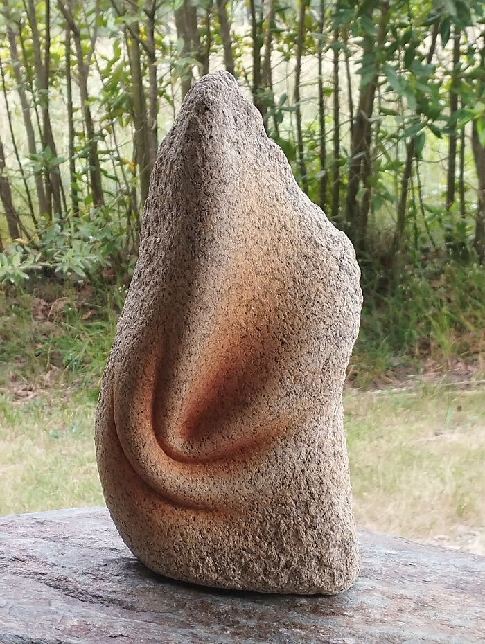 A smooth, soft-looking stone sculpture with a textured surface, displayed outdoors with trees in the background.