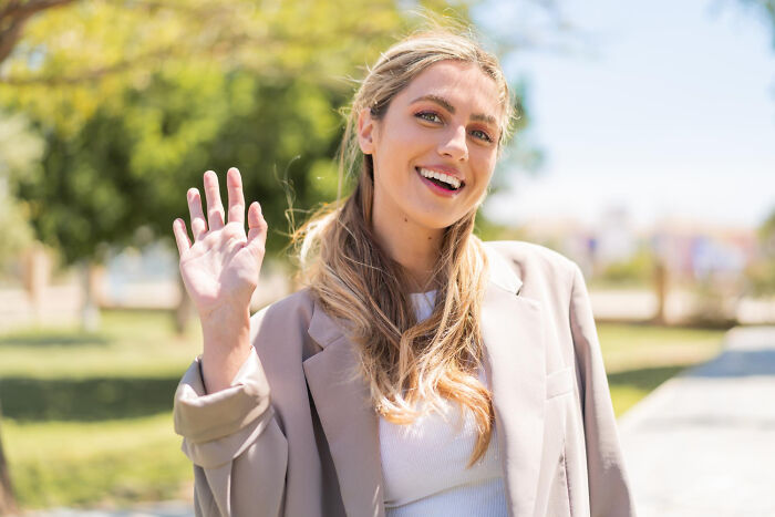 Woman waving in a park, wearing a beige blazer, smiling on a sunny day.