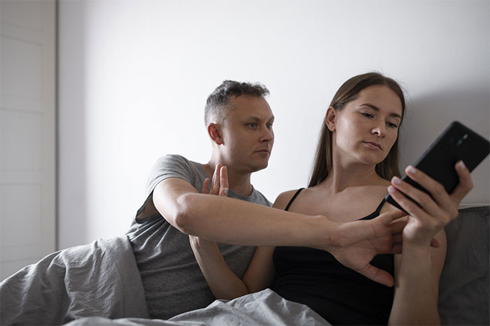Couple in bed with woman checking phone, man appearing curious or concerned.