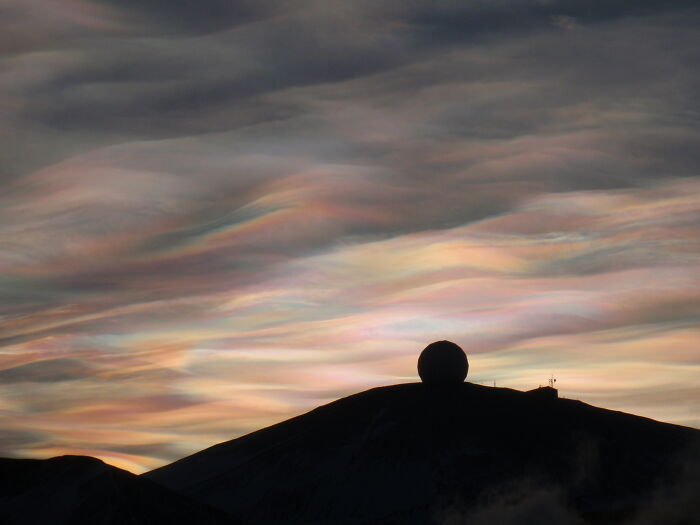 Colorful iridescent clouds above a silhouetted hilltop, showcasing stunning natural phenomena.