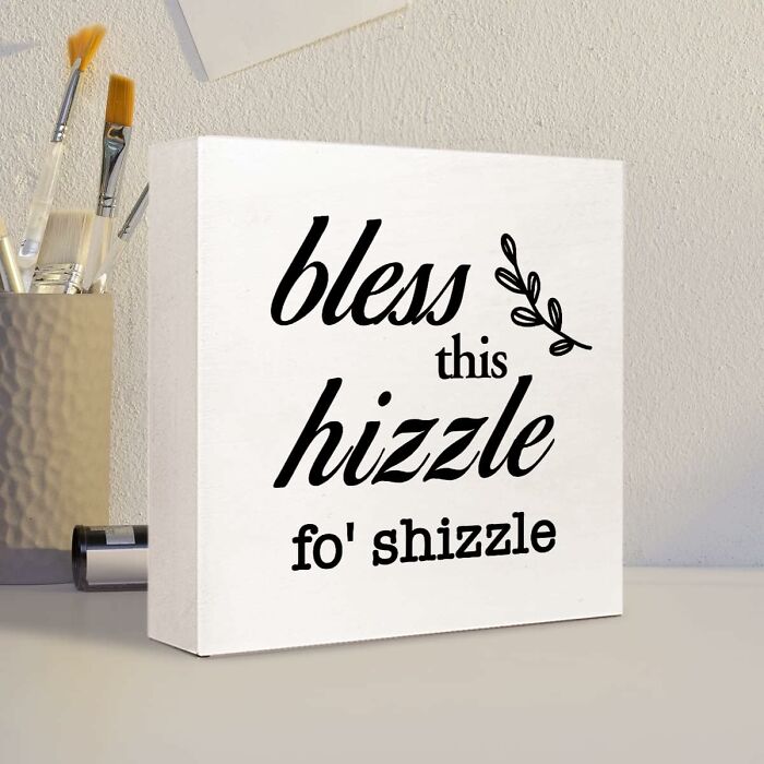Funny Amazon find: Decorative sign reading "bless this hizzle fo' shizzle" on a desk beside paintbrushes.