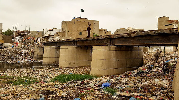 Urban hell scene with a bridge over a trash-filled river, surrounded by rundown buildings and debris.