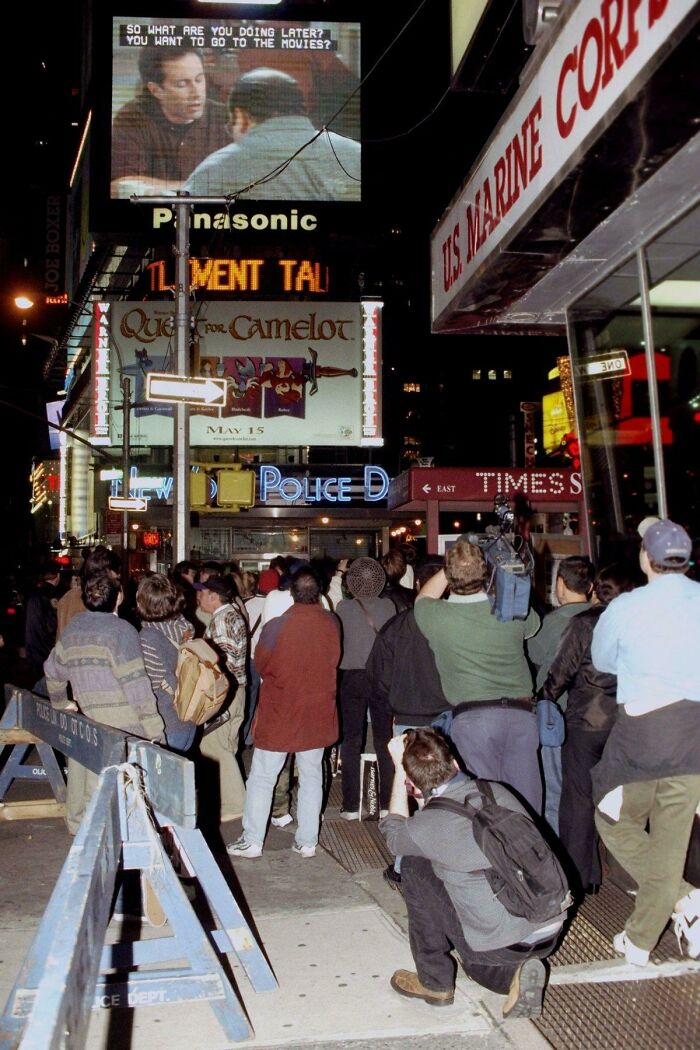Crowd watching a large screen in Times Square at night displaying a historical TV moment.