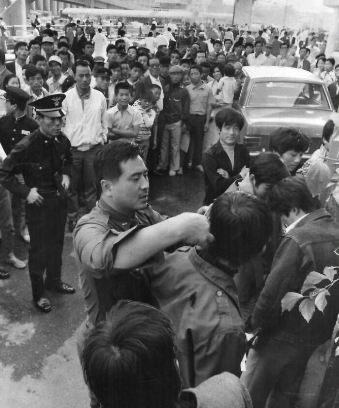 Crowd gathered in a historical photo, a police officer inspects a man's collar in an orderly yet tense scene.