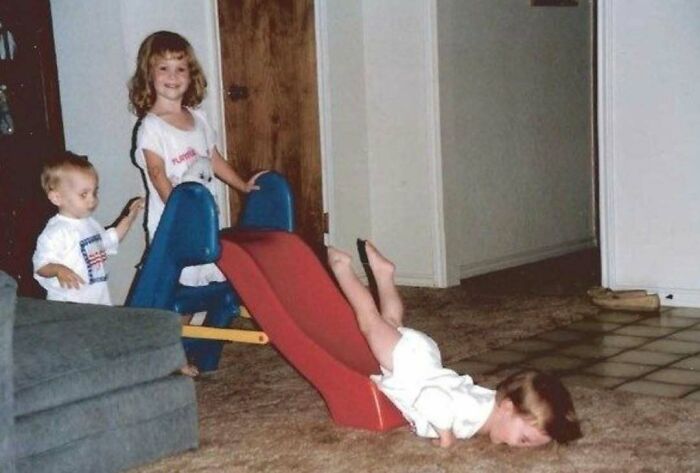 Awkward family photo of children playing on a slide indoors, with one child sliding headfirst onto the carpet.