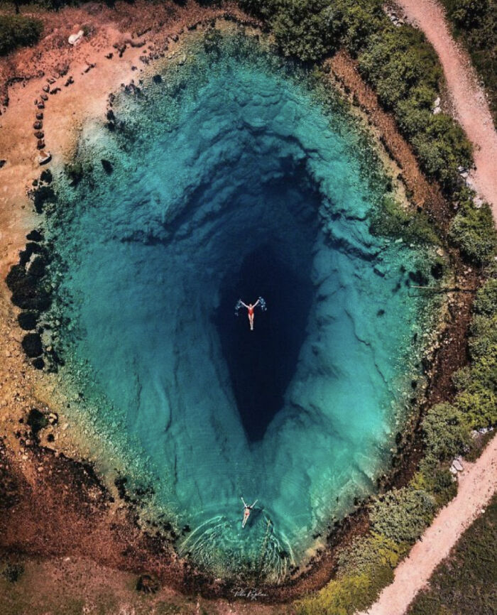 Aerial view of a massive natural sinkhole with swimmers in clear blue water, illustrating megafobia and scarily big formations.