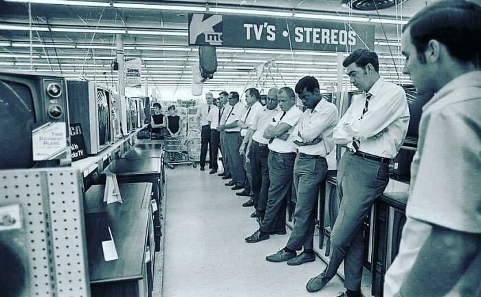 Men watching TV displays in a 1960s store under a vintage electronics sign. Fascinating historical pic.