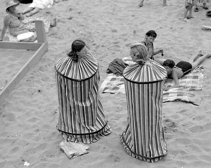Two women in vintage beach changing tents on a sandy beach, capturing a fascinating historical moment.