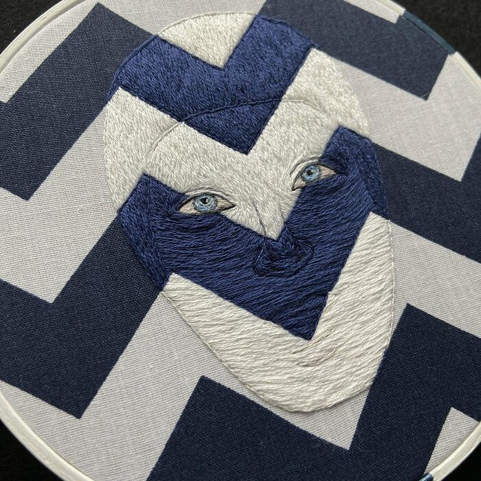 Abstract face embroidery design with intricate stitches in blue and white on chevron fabric background.