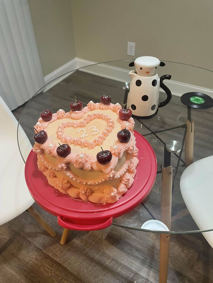 Heart-shaped cake with pink frosting and cherries, placed on a red platter. Delightful bakers' creation on a glass table.