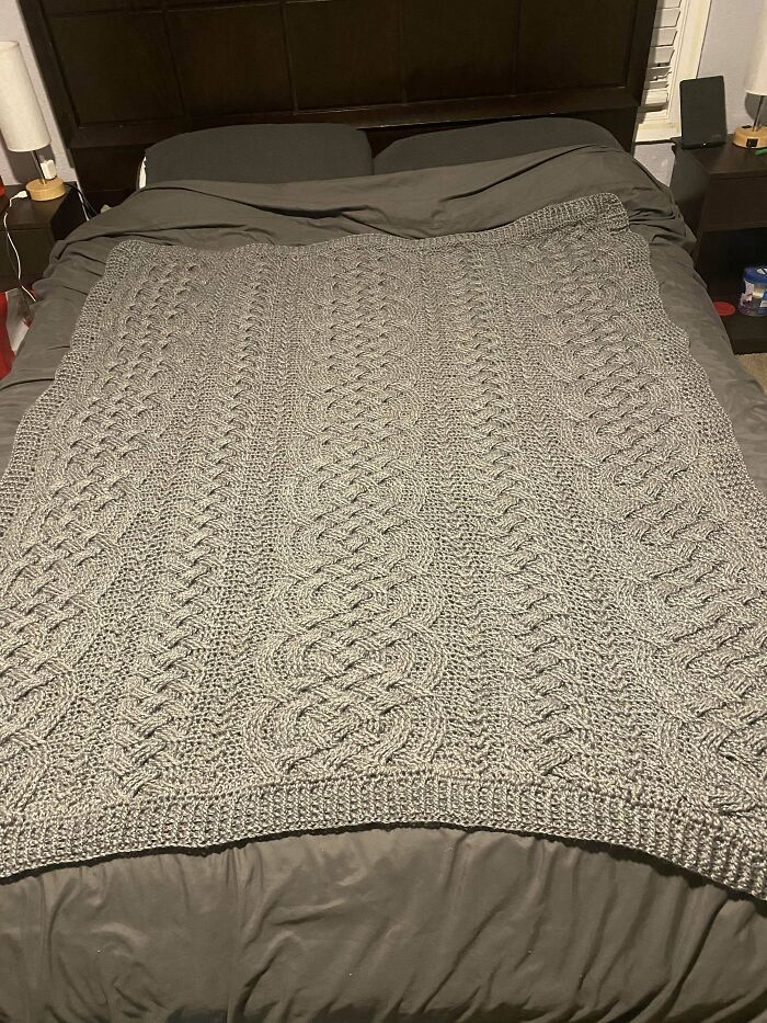 Crochet enthusiasts proudly display a large, intricately patterned gray crochet blanket spread out on a bed.