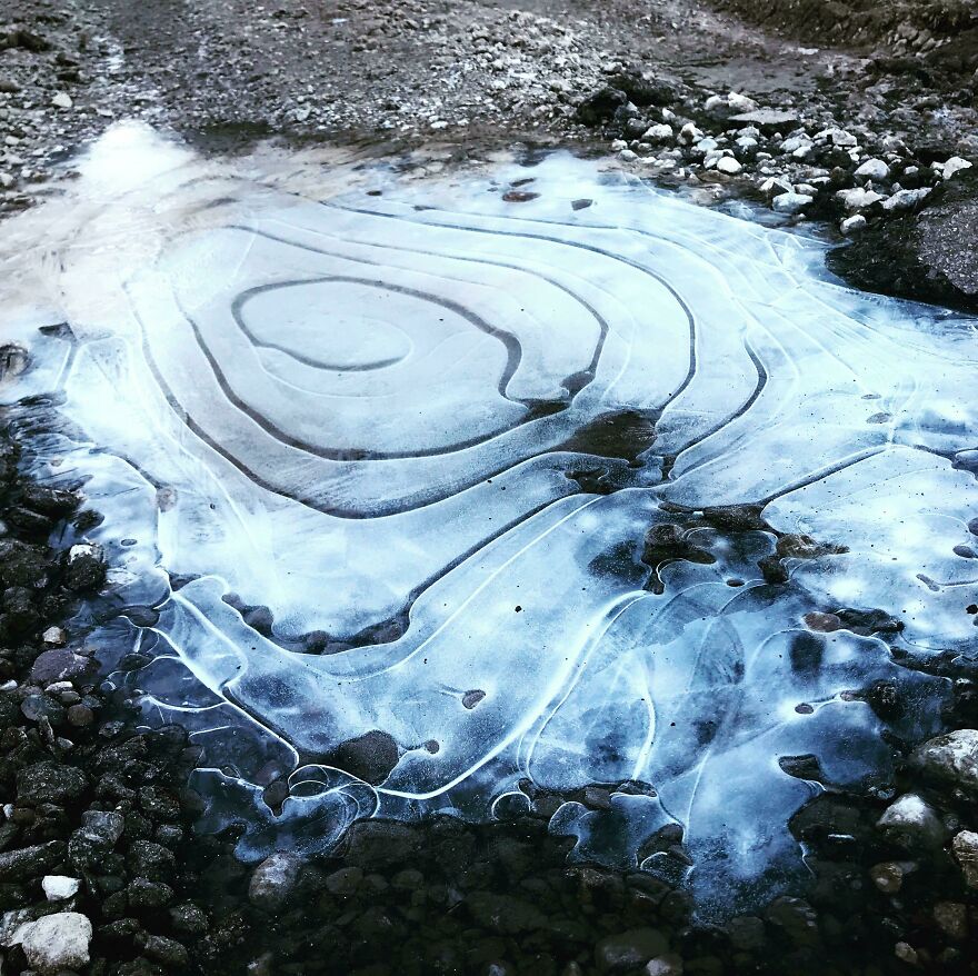Frozen puddle resembling accidental art with intricate patterns, surrounded by gravel and earth.