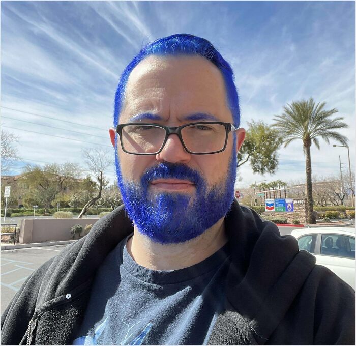 Man with bright blue hair and beard, wearing glasses and a dark shirt, standing outdoors under a clear sky. Changing-Hair-Color.