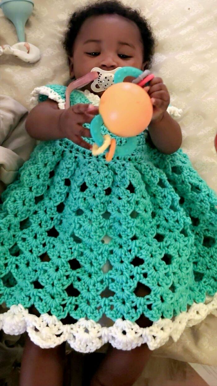 Baby wearing a handmade crochet dress in teal and white, holding a colorful toy while lying on a bed.
