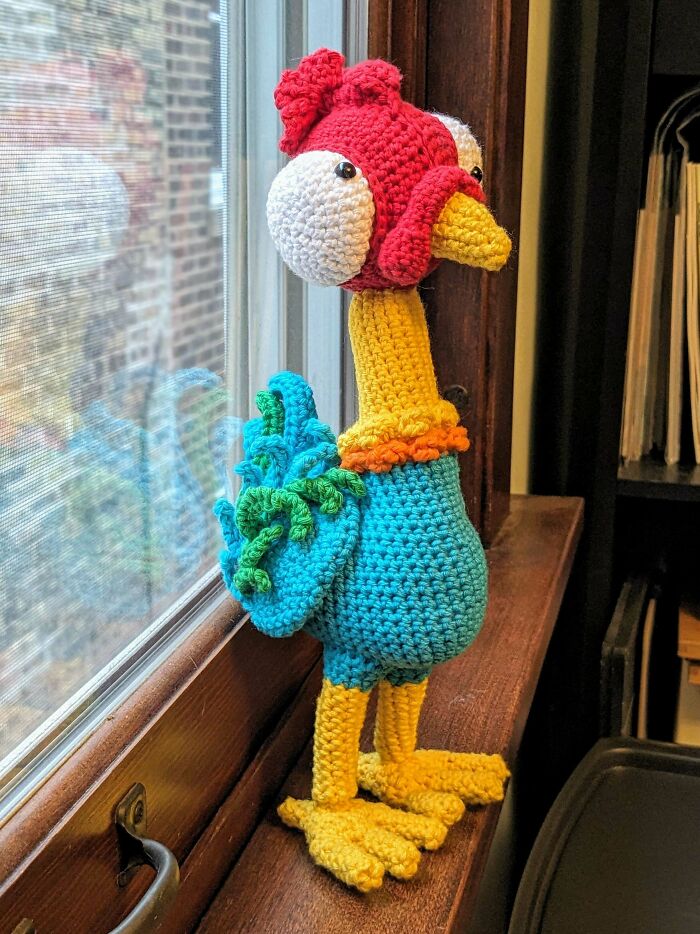 Crochet enthusiast’s colorful rooster amigurumi with big eyes, standing on a wooden windowsill indoors.